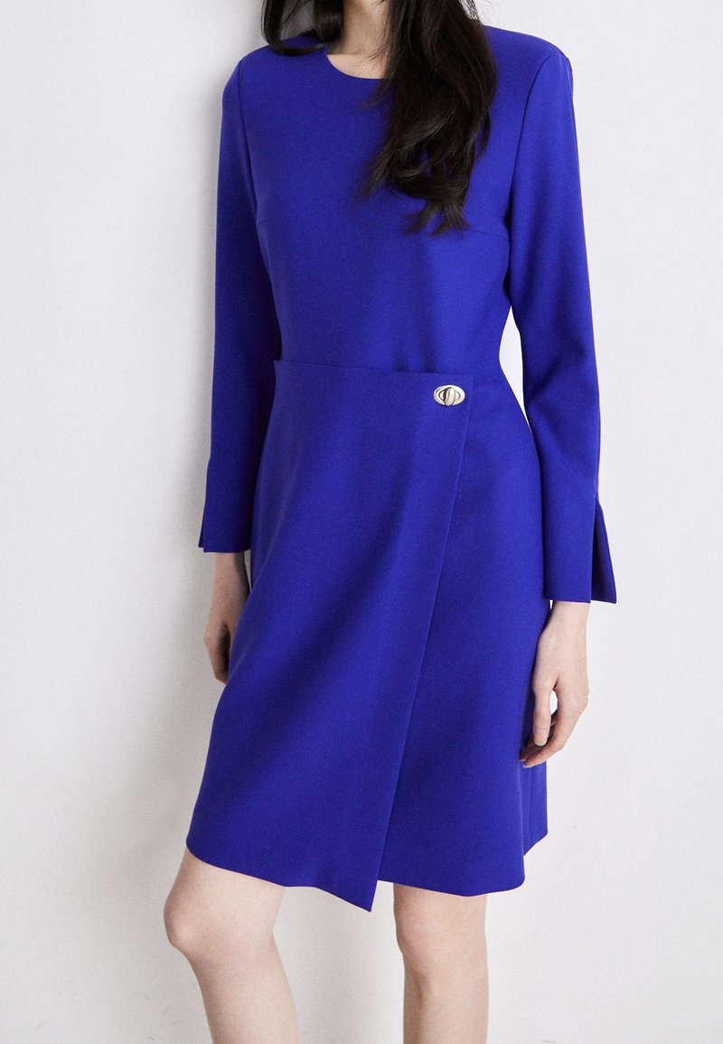 Femme portant une robe bleu roi, longue jusqu'aux genoux, à manches longues, avec un rabat asymétrique à l'avant fermé par un petit bouton doré.