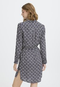 Moteris su pečių ilgio plaukais vilki ilgas rankoves turintį su diržu suknelę su juodai balta geometrine raštu, stovinti prieš paprastą foną.