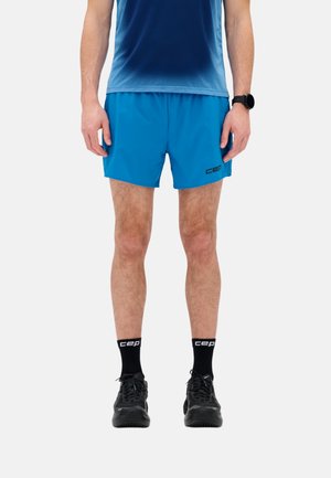 Male wearing blue athletic shorts and shirt, black socks, black running shoes, and a black wristwatch, standing against a white background.