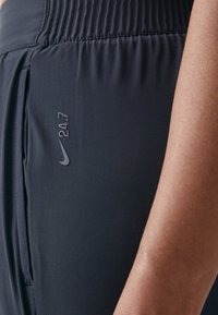 Črne atletske hlače iz gladkega materiala, z nabranim pasom in diskretnim logotipom Nike ter napisom "24.7" na strani.