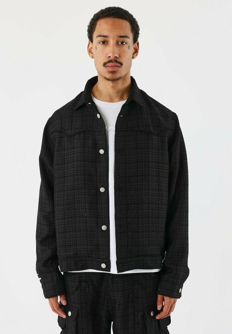 Veste noire à boutons, fabriquée en tissu texturé, présentant un motif, un col classique et des détails en métal argenté. Portée par-dessus une chemise blanche.