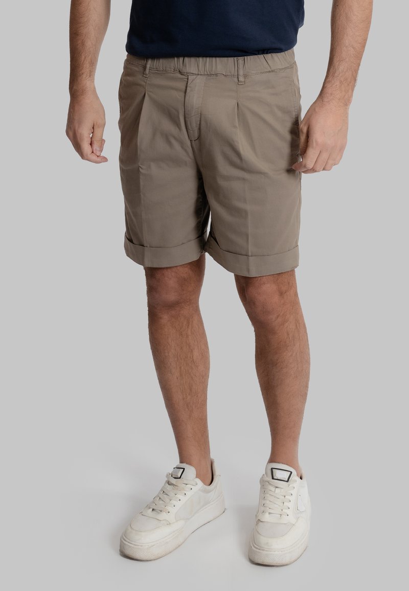 Pantaloni corti beige con cintura elastica, davanti plissettato, orli arrotolati e una texture liscia, abbinati a sneaker bianche.