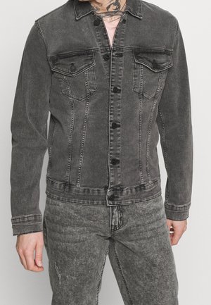 Homme portant une veste en jean noire délavée foncée et un jean assorti, avec un tatouage visible sur le torse et une chemise rose clair en dessous.