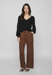 Suéter negro de cuello en V con mangas largas, combinado con pantalones de pierna ancha marrones, que tienen una textura suave y bolsillos frontales. Tacones abiertos negros.