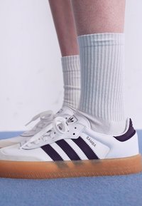 White leather sneakers with purple stripes, gum sole, and round laces. Worn with ribbed white socks. Minimalistic design, athletic style.