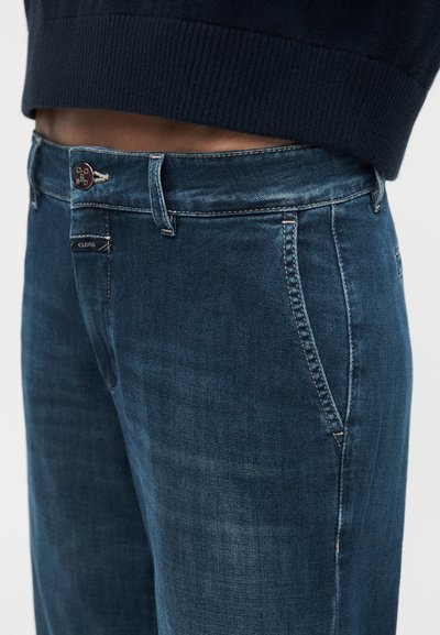 Jean en denim bleu foncé avec poches avant et un bouton de marque, porté avec un pull court sombre laissant apparaître la ligne de taille.