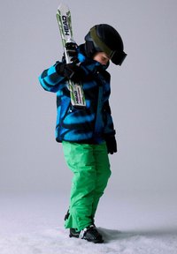 Tenue de ski avec une veste à motifs bleus et noirs, un pantalon vert, des gants noirs et un casque, tenant un ski blanc avec des accents noirs, sur la neige.