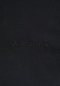 Čierna rebrovaná tkanina s embosovaným textovým dizajnom, ktorý znie "9N1M SENSE". Žiadne ďalšie vzory ani kovové súčiastky nie sú viditeľné.