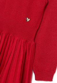 Rotes Strickkleid mit einem gerippten Oberteil und einem ausgestellten Rock. Verfügt über ein glänzendes, goldenes Herz-Akzent auf der Brust. Weiche Textur mit leichtem Schimmer.