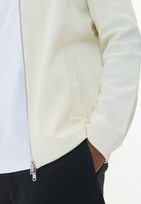 Svetlo kremna zip-up jakna z teksturirano tkanino in pletenimi manšetami, v kombinaciji z belo majico in črnimi hlačami. Ima stranske žepe in dvojne zadrge.