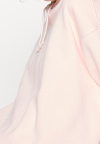 Sudadera oversized de color rosa claro con capucha ajustable, textura de tela suave y diseño simple, que presenta un detalle en la costura lateral.