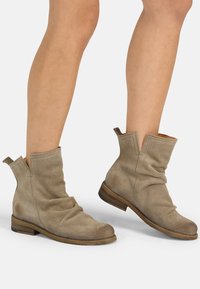 Botas de tobillo beige hechas de cuero perforado con un diseño arrugado, tacón bajo de madera y lengüetas para facilitar su uso.