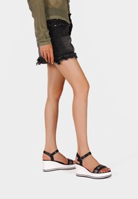 Sandali platform neri con fasce larghe e fibbia alla caviglia, dotati di una suola a zeppa bianca con dettagli neri. Indossati con una gonna di jeans strappata.