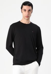 Schwarzes Langarmshirt aus glattem Material mit Rundhalsausschnitt und kleinem Logo auf der Brust. Das Modell trägt weiße Hosen.