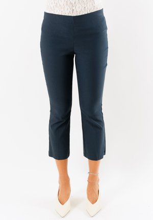 Donna che indossa pantaloni corti blu navy, tacchi alti bianchi e un top di pizzo bianco, in piedi su uno sfondo bianco uniforme.