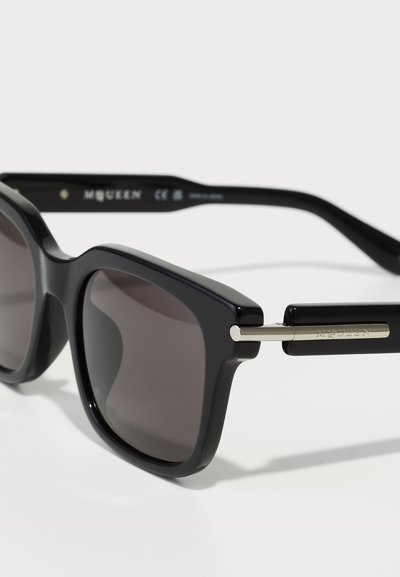 Ochelari de soare negri cu un cadru pătrat îndrăzneț, lentile întunecate și accente metalice pe ochelari, având inscripționat "MCQUEEN".