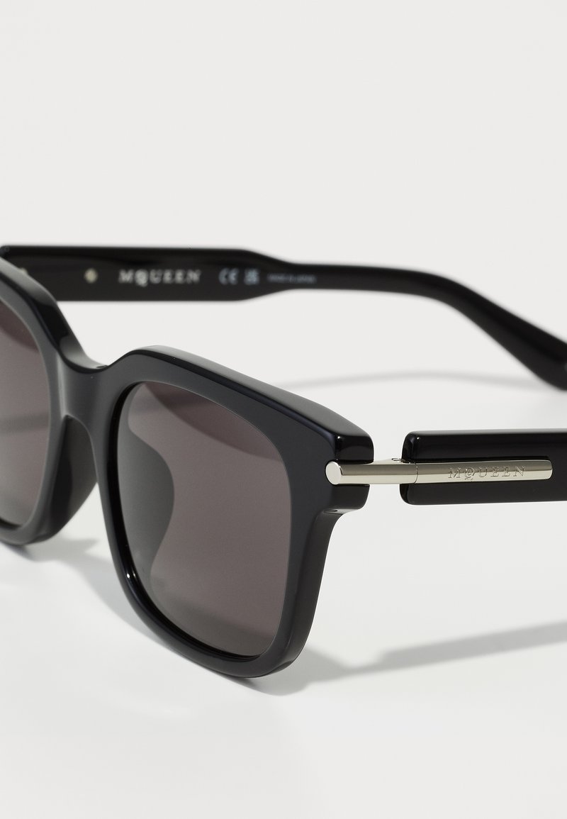 Lunettes de soleil noires avec une monture carrée audacieuse, des verres teintés foncés et des accents métalliques sur les branches avec l'inscription "MCQUEEN" gravée.