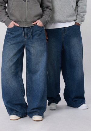 Twee mensen die oversized blauwe spijkerbroeken, grijze hoodies en witte sneakers dragen, naast elkaar staan met handen in de zakken.