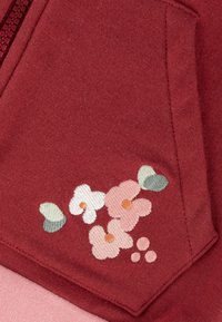 Rote Fleece-Haube mit einer Seitentasche, die bestickte Blumen in Pink, Weiß und Grün zeigt, mit weicher Textur und detaillierter Naht.