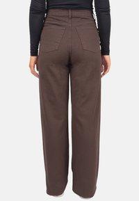 Pantalon marron taille haute à jambes larges, avec des poches arrière et un tissu texturé. Porté avec un haut noir ajusté à manches longues.