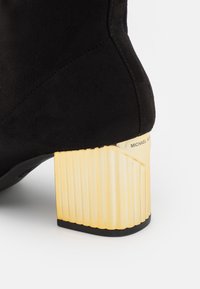 Bottine en suède noir avec talon bloc en or côtelé, arborant la marque "Michael Kors" près du bord supérieur.