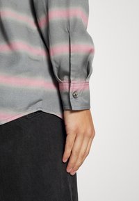 Camisa gris con rayas rosas, puño abotonado y textura suave. El dobladillo es ligeramente curvado, combinado con mezclilla oscura.