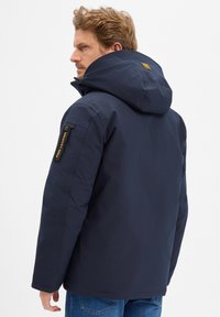 Marineblauwe jas met een capuchon, voorzien van een met rits afsluitbare zak op de linker mouw en een logo-detail. Heeft een gladde textuur en een reguliere pasvorm.