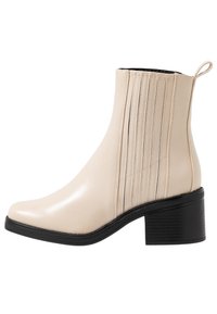 Bottine synthétique beige avec une finition lisse, un bout arrondi et des coutures verticales. Elle dispose d'un talon bloc noir et d'une languette pour un enfilage facile.