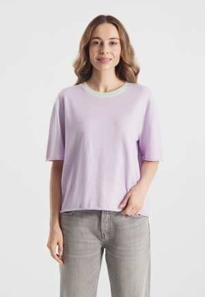 Vrouw met golvend bruin haar en een lichtpaarse trui met korte mouwen en grijze spijkerbroek, staand tegen een witte achtergrond.