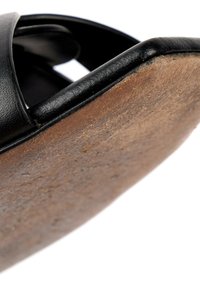 Gros plan d'une sandale montrant une bride en cuir noir et une semelle marron usée sur fond blanc.