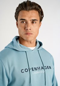 Sudadera de color azul claro con un bolsillo canguro frontal, capucha con cordón y logos impresos de "COPENHAGUEN" y "LINDBERGH" en colores más oscuros. Textura suave.