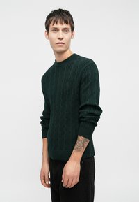 Σακάκι καλωδίων σε σκούρο πράσινο, με στρογγυλή λαιμόκοψη και ριμπ μανσέτες. Η υφή είναι ανάγλυφη, με εφαρμοστή σιλουέτα και μακριά μανίκια.