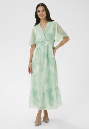 Femme souriante, portant une robe portefeuille tie-dye vert clair avec des manches volantées et un ourlet à volants, des chaussures pointues beiges, debout avec la main sur la hanche.