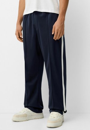 Mężczyzna w granatowych spodniach dresowych z białymi paskami po bokach i białych trampkach, stojący na jednolitym białym tle.