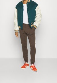 Turkos och krämfärgad collegejacka med ribbad krage, bärs över en vit t-shirt, parat med bruna byxor och orangea sneakers.