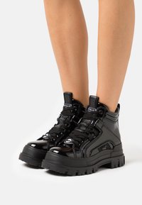 Zapatillas altas de charol negro con suelas gruesas de goma, sujetas con cordones en la parte delantera y acentos de logotipo. Textura suave y diseño voluminoso.