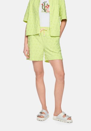 Lime green textured shorts with drawstring, matching button-up shirt, white cactus print T-shirt, and white sandals with jeweled buckles.