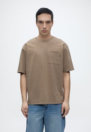 Homme aux cheveux courts et noirs portant un t-shirt marron ample avec une poche poitrine et un jean bleu clair, debout devant un fond blanc uni.