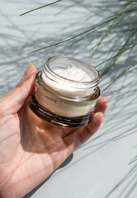 Korres White Pine volymgivande serum-i-fuktkräm i en klar glasburk med en blank guldbas, med en texturerad krämig inredning och etiketter.