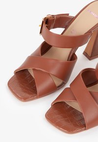Brune læder høje hæle sandaler med firkantet tå, krydsende remme og et tekstureret krokodillemønster på sålen. Guld spænde detalje inkluderet.