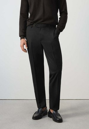 Hombre con pantalones de vestir negros, suéter marrón oscuro de manga larga y mocasines de cuero negro, de pie sobre un suelo gris.