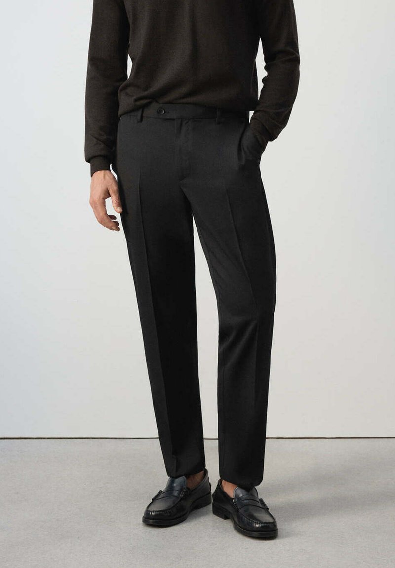 Homme portant un pantalon habillé noir, un pull à manches longues marron foncé et des mocassins en cuir noir, debout sur un sol gris.