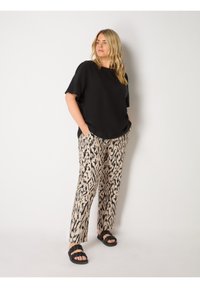 Svart kortärmad topp i kombination med beige byxor med ett utmärkande svart abstrakt mönster. Modellen bär svarta sandaler.