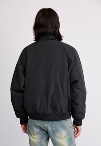 Veste bombers noire avec un col et des poignets côtelés, présentant une texture lisse. Associée à un jean bleu clair délavé.