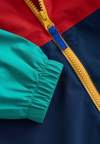 Veste colorée avec des panneaux rouges, turquoise et bleu marine. Fermeture éclair jaune avec un accent bleu. Texture lisse avec des extrémités de manches froncées.
