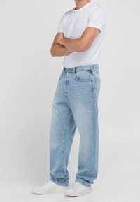 Man som bär ljusblå jeans, vit t-shirt och vita sneakers, stående med armarna i kors mot en enfärgad bakgrund.