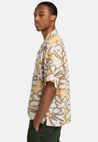 Joven con pantalones verdes que lleva una camisa de manga corta de color beige y amarillo con un diseño floral, de pie de perfil con las manos en los bolsillos.
