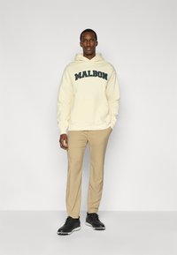 Κρεμ φούτερ με κουκούλα που φέρει τη λέξη "MALBON" με σκούρα πράσινα γράμματα, συνδυασμένο με μπεζ παντελόνι και μαύρα παπούτσια. Το ύφασμα φαίνεται μαλακό με άνετη εφαρμογή.