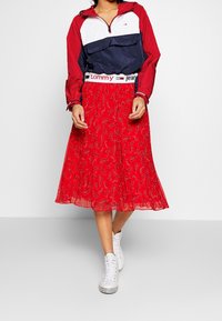 Röd midi-skjort med paisley-mönster och en genomskinlig textur, kombinerad med en marinblå och vit zip-up jacka och vita sneakers.