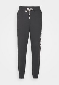 Zwarte joggingsbroek met witte trekkoord, elastische boorden en witte grafische tekst die verticaal op het rechterbeen loopt.